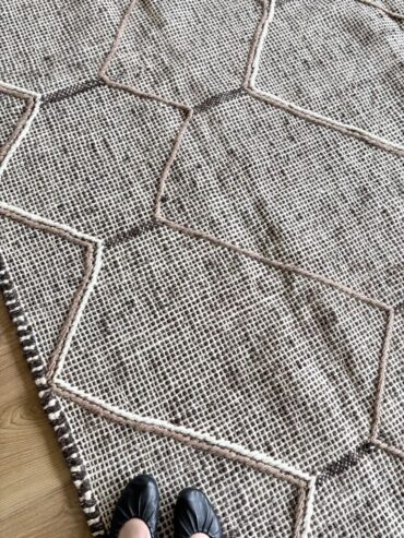Alfombra kilim artesanal de lana en tonos crudo y negro con dibujo geométrico en relieve, tejida a mano en Marruecos, sobre suelo de madera clara.